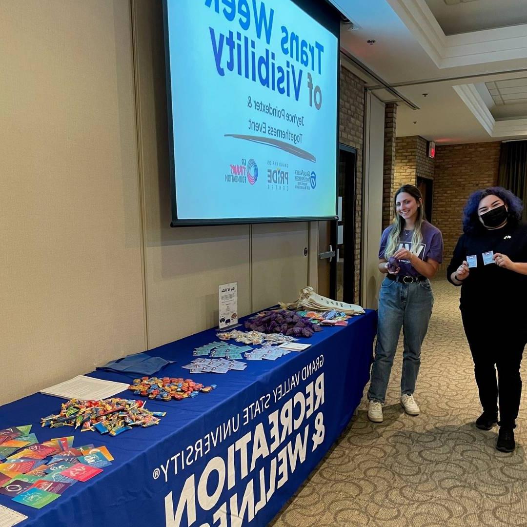 Image of two RecWell staff by a table full of fun wellness supplies in front of "Trans Week of Visibility" projected on a screen